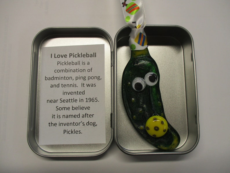 Peut inclure: Une petite d&eacute;coration en verre en forme de cornichon vert avec des yeux globuleux et un charme de balle de pickleball jaune. La d&eacute;coration est dans une bo&icirc;te en &eacute;tain argent&eacute;e avec une carte blanche qui dit "J'aime le pickleball. Le pickleball est une combinaison de badminton, de ping-pong et de tennis. Il a &eacute;t&eacute; invent&eacute; pr&egrave;s de Seattle en 1965. Certains pensent qu'il porte le nom du chien de l'inventeur, Pickles."