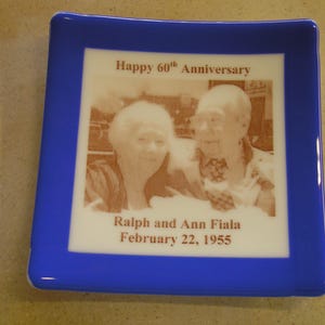 May include: A blue glass plaque with a white border and a photo of an older couple. The text reads "Happy 60th Anniversary" and "Ralph and Ann Fiala February 22, 1955".
