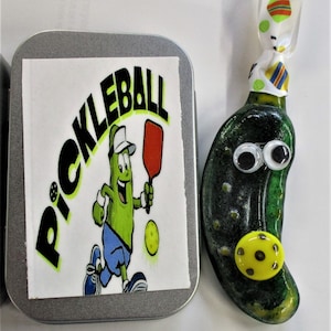 Peut inclure: Une d&eacute;coration en forme de cornichon vert avec des yeux globuleux et une balle de pickleball jaune. La d&eacute;coration est attach&eacute;e &agrave; un ruban blanc &agrave; pois color&eacute;s. Une bo&icirc;te en &eacute;tain avec un autocollant blanc qui dit "Pickleball" est &eacute;galement dans l'image.
