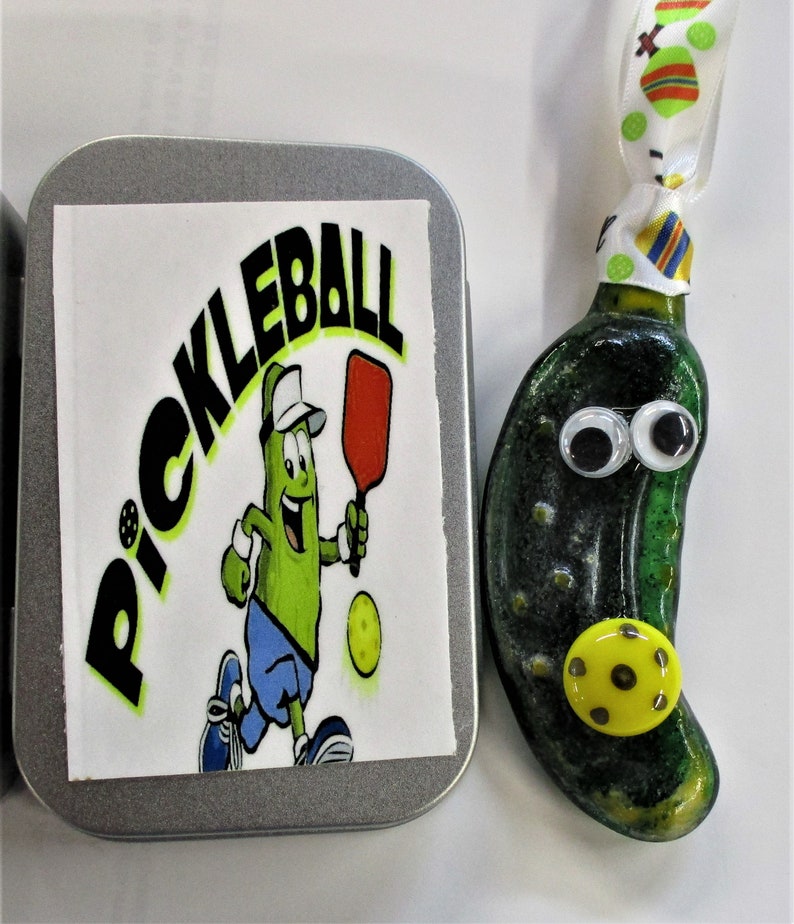 Peut inclure: Une d&eacute;coration en forme de cornichon vert avec des yeux globuleux et une balle de pickleball jaune. La d&eacute;coration est attach&eacute;e &agrave; un ruban blanc &agrave; pois color&eacute;s. Une bo&icirc;te en &eacute;tain avec un autocollant blanc qui dit "Pickleball" est &eacute;galement dans l'image.