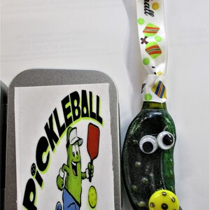 Peut inclure: Une d&eacute;coration en forme de cornichon vert avec des yeux globuleux et une balle de pickleball jaune. La d&eacute;coration est attach&eacute;e &agrave; un ruban blanc sur lequel est imprim&eacute; le mot "Pickleball". La d&eacute;coration est dans une bo&icirc;te en argent avec un autocollant qui dit "Pickleball" et repr&eacute;sente un cornichon de dessin anim&eacute; jouant au pickleball.