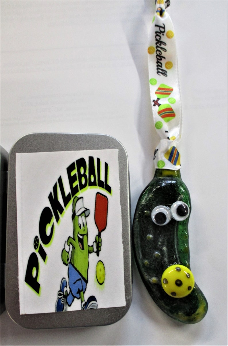 Peut inclure: Une d&eacute;coration en forme de cornichon vert avec des yeux globuleux et une balle de pickleball jaune. La d&eacute;coration est attach&eacute;e &agrave; un ruban blanc sur lequel est imprim&eacute; le mot "Pickleball". La d&eacute;coration est dans une bo&icirc;te en argent avec un autocollant qui dit "Pickleball" et repr&eacute;sente un cornichon de dessin anim&eacute; jouant au pickleball.
