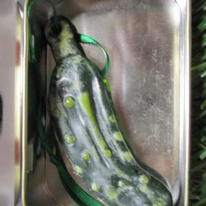 May include: A green pickle-shaped Christmas ornament with a green ribbon, in a silver tin.