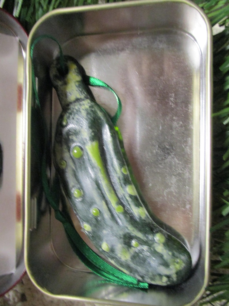 May include: A green pickle-shaped Christmas ornament with a green ribbon, in a silver tin.
