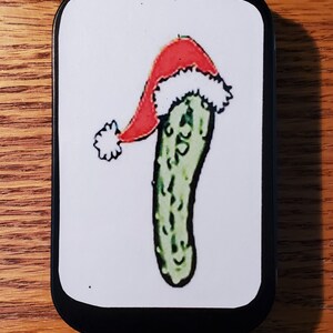 May include: A green pickle wearing a red and white Santa hat. The pickle is on a white background inside a black tin.