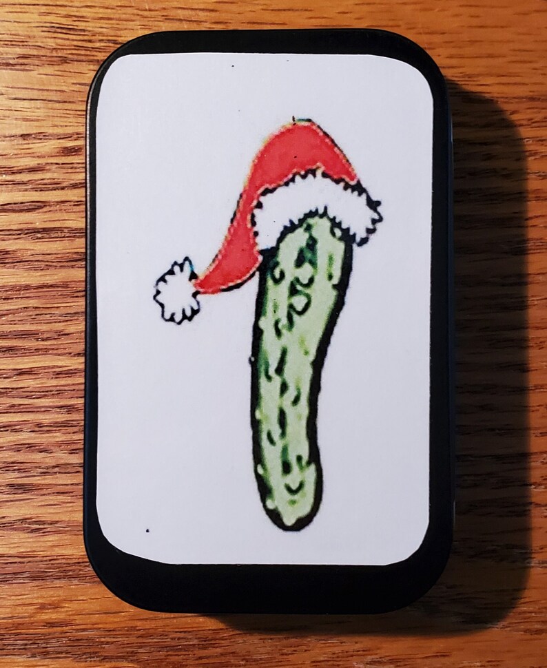 May include: A green pickle wearing a red and white Santa hat. The pickle is on a white background inside a black tin.