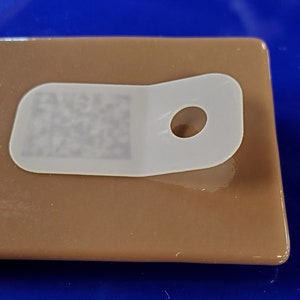 May include: A white plastic tag with a hole in the center and a barcode on the left side. The tag is on a brown surface.