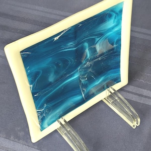 May include: A blue and white glass art piece with a white frame and clear acrylic stand.
