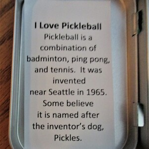 Peut inclure: Une bo&icirc;te en &eacute;tain argent&eacute;e avec une carte blanche &agrave; l'int&eacute;rieur. La carte contient le texte suivant : "J'aime le pickleball. Le pickleball est une combinaison de badminton, de ping-pong et de tennis. Il a &eacute;t&eacute; invent&eacute; pr&egrave;s de Seattle en 1965. Certains pensent qu'il porte le nom du chien de l'inventeur, Pickles."