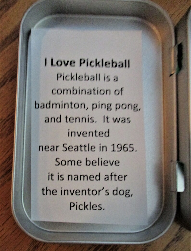Peut inclure: Une bo&icirc;te en &eacute;tain argent&eacute;e avec une carte blanche &agrave; l'int&eacute;rieur. La carte contient le texte suivant : "J'aime le pickleball. Le pickleball est une combinaison de badminton, de ping-pong et de tennis. Il a &eacute;t&eacute; invent&eacute; pr&egrave;s de Seattle en 1965. Certains pensent qu'il porte le nom du chien de l'inventeur, Pickles."