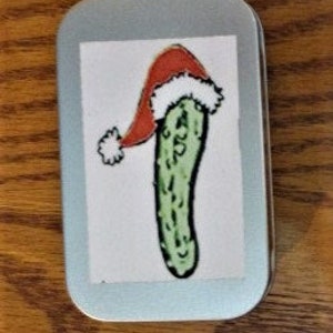 May include: A small, silver tin with a green pickle wearing a red Santa hat. The pickle is green with white bumps and the hat has a white pom-pom.