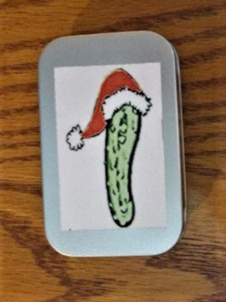May include: A small, silver tin with a green pickle wearing a red Santa hat. The pickle is green with white bumps and the hat has a white pom-pom.