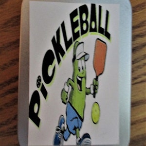 Peut inclure: Une bo&icirc;te en &eacute;tain argent&eacute;e avec un autocollant blanc repr&eacute;sentant un personnage de cornichon de dessin anim&eacute; tenant une pagaie et une balle. L'autocollant dit "Pickleball" en lettres noires.
