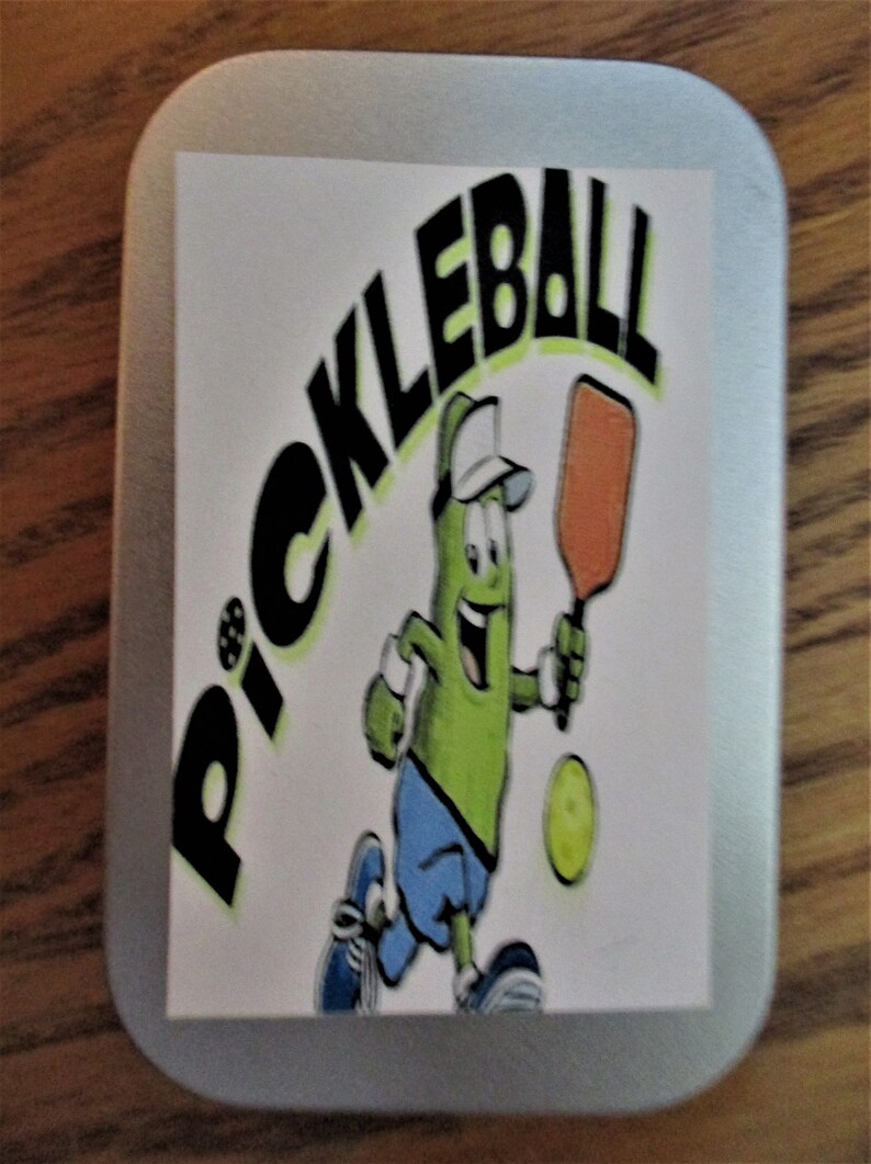 Peut inclure: Une bo&icirc;te en &eacute;tain argent&eacute;e avec un autocollant blanc repr&eacute;sentant un personnage de cornichon de dessin anim&eacute; tenant une pagaie et une balle. L'autocollant dit "Pickleball" en lettres noires.