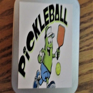 Peut inclure: Un carr&eacute; blanc aux coins arrondis pr&eacute;sente un cornichon de dessin anim&eacute; jouant au pickleball. Le cornichon porte une chemise bleue, un chapeau blanc et tient une raquette. Le mot "PICKLEBALL" est &eacute;crit en lettres noires au-dessus du cornichon.