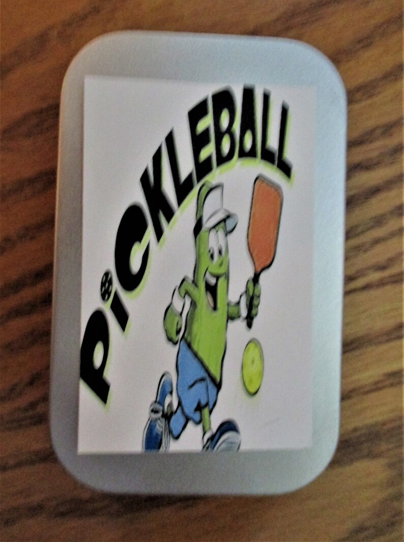 Peut inclure: Un carr&eacute; blanc aux coins arrondis pr&eacute;sente un cornichon de dessin anim&eacute; jouant au pickleball. Le cornichon porte une chemise bleue, un chapeau blanc et tient une raquette. Le mot "PICKLEBALL" est &eacute;crit en lettres noires au-dessus du cornichon.