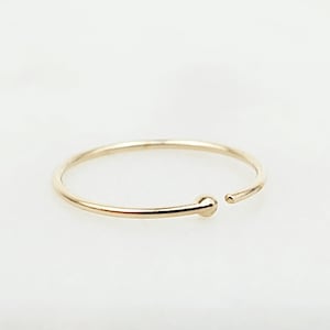 May include: A delicate gold nose ring with a small ball detail. The ring is open-ended and sits on a white marble surface. The ring is a simple, elegant piece of jewelry.