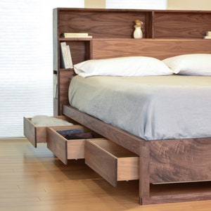May include: A wooden bed frame with a headboard that has built-in shelves. The bed has three drawers that are pulled out, revealing the interior.