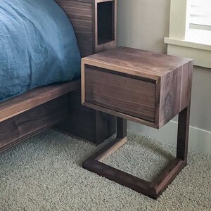 May include: A dark brown wooden nightstand with a single drawer and a square base. The nightstand is made of walnut wood and has a modern design.