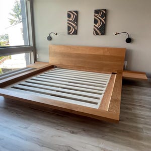 May include: A modern, light-colored wooden bed frame with a headboard and integrated side tables. The bed frame has a low profile design and a slatted base. The room has large windows and decorative wall art.