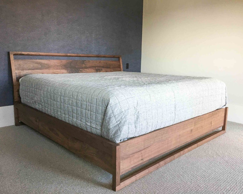 Puede incluir: Un marco de cama de madera con un edred&oacute;n acolchado gris claro. El marco de la cama tiene un dise&ntilde;o simple y moderno con una cabecera baja y un piecero ligeramente elevado.