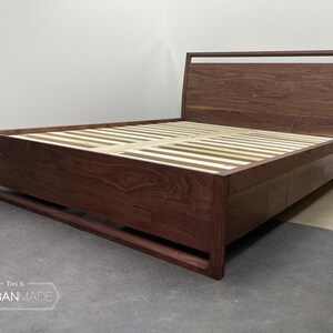 Puede incluir: Un marco de cama plataforma de madera oscura con una cabecera de listones y un caj&oacute;n de almacenamiento debajo. El marco de la cama est&aacute; hecho de madera maciza y tiene un acabado natural.