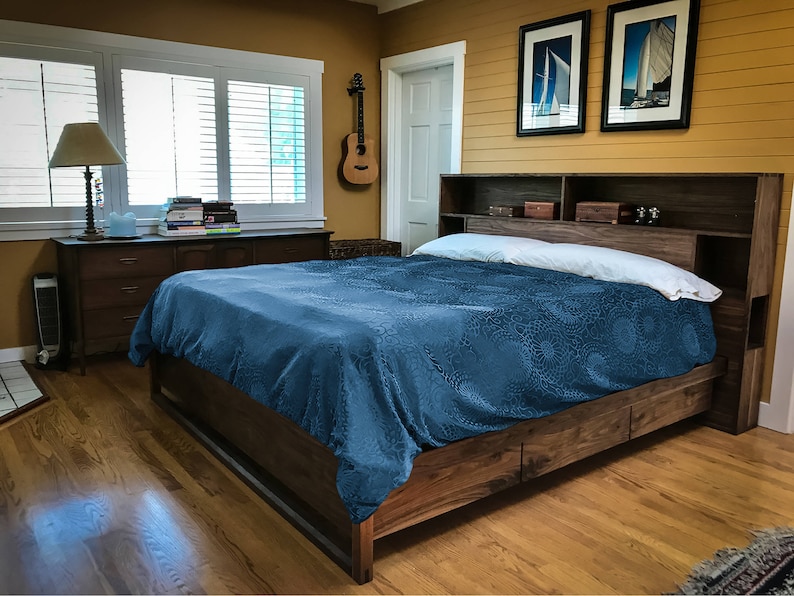 May include: A wooden bed frame with a headboard that includes built-in shelves. The bed has a blue patterned duvet and a white pillow. The bed is in a bedroom with wood floors and a dresser with books on top.
