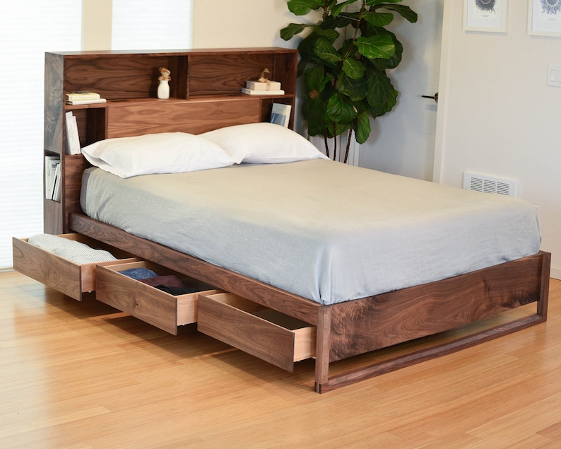 May include: A wooden bed frame with a headboard that has built-in shelves. The bed has three drawers that pull out from the bottom. The bed is made of walnut wood and has a light gray mattress.
