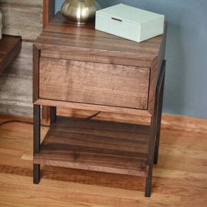 May include: A dark brown wooden nightstand with a drawer and a lower shelf. The nightstand has a metal frame and a light green box on top. The wood grain is visible. A silver-colored lamp sits on top.