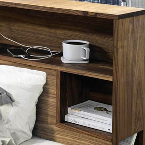 May include: A wooden headboard with built-in shelves and a charging station. The headboard has a light brown finish and features two shelves, one above the other. The top shelf has a white coffee mug and a phone on it. The bottom shelf has a stack of books. The charging station has two outlets and two USB ports.