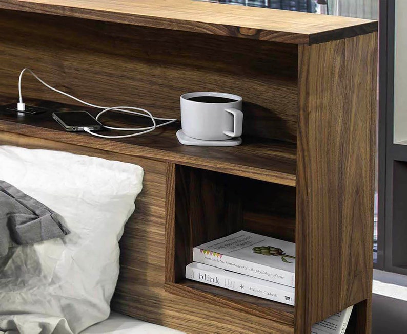 May include: A wooden headboard with built-in shelves and a charging station. The headboard has a light brown finish and features two shelves, one above the other. The top shelf has a white coffee mug and a phone on it. The bottom shelf has a stack of books. The charging station has two outlets and two USB ports.