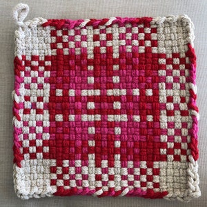 May include: Square, woven hot pad in shades of red, pink, and off-white. The center features a heart-shaped design, with a loop for hanging. The edges are finished with a braided red trim. A handmade kitchen accessory.