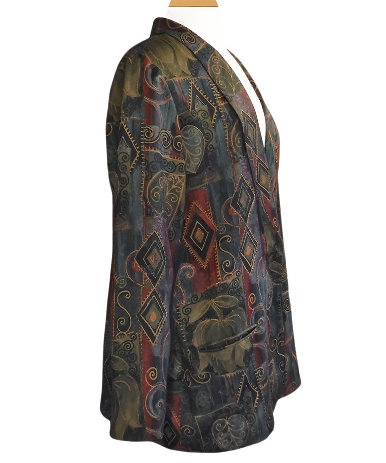 May include: A patterned blazer in shades of green, burgundy, and black. The blazer features a geometric design with diamond shapes and floral accents. It has a notched collar and two front pockets. The blazer is displayed on a mannequin.