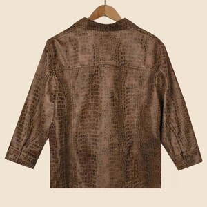 May include: A brown, button-down shirt with a faux crocodile skin texture. The shirt has three-quarter sleeves and a classic collar. The back of the shirt is visible, hanging on a wooden hanger.