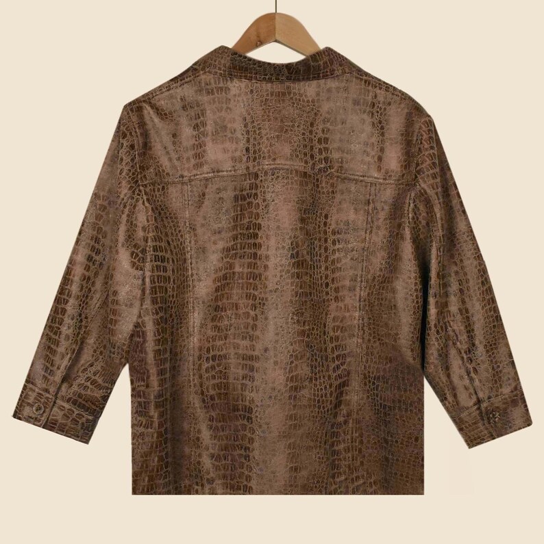 May include: A brown, button-down shirt with a faux crocodile skin texture. The shirt has three-quarter sleeves and a classic collar. The back of the shirt is visible, hanging on a wooden hanger.