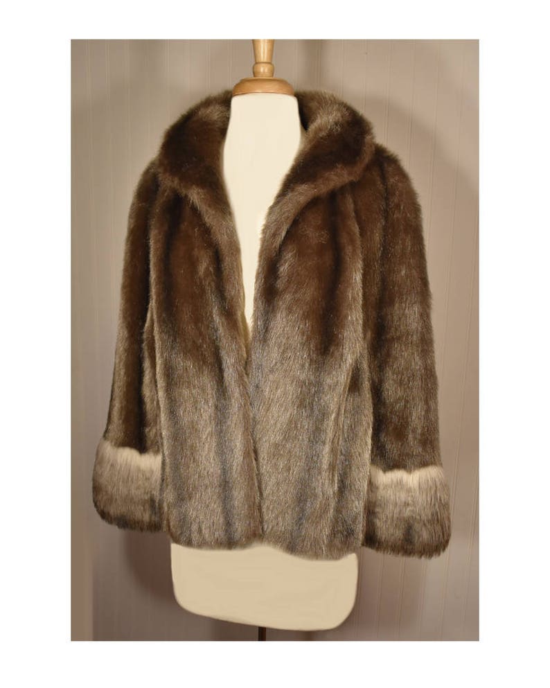 Peut inclure: Une veste en fausse fourrure marron avec un col ch&acirc;le et des manches longues. Les poignets sont bord&eacute;s d'une fourrure plus claire.