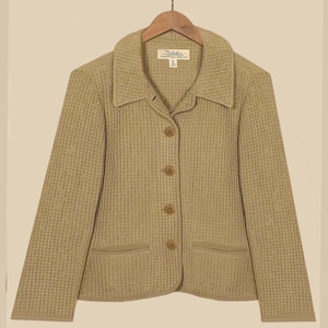 Puede incluir: Chaqueta beige con tejido texturizado, cuello, cuatro botones y dos bolsillos delanteros. La chaqueta está colgada de una percha de madera. La etiqueta dice "Cabela's".
