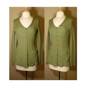 Könnte beinhalten: Ein langärmliger, olivgrüner Cardigan mit Knopfleiste und zwei Taschen. Der Cardigan ist aus geripptem Stoff und hat eine leicht taillierte Silhouette.