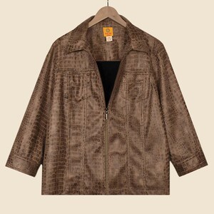 May include: A brown faux-leather jacket with a crocodile skin texture. The jacket features a collared neckline, two chest pockets, and a full-length zip. The sleeves are three-quarter length. The jacket hangs on a wooden hanger.