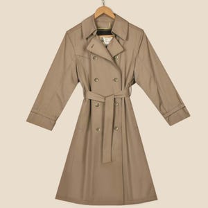 K&ouml;nnte beinhalten: Ein beiger, zweireihiger Trenchcoat mit G&uuml;rtel und langen &Auml;rmeln. Der Mantel hat ein klassisches Design und ist aus einem leichten Stoff gefertigt.