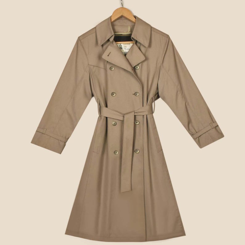K&ouml;nnte beinhalten: Ein beiger, zweireihiger Trenchcoat mit G&uuml;rtel und langen &Auml;rmeln. Der Mantel hat ein klassisches Design und ist aus einem leichten Stoff gefertigt.