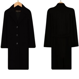 May include: A black long-sleeved coat with a collar and three buttons down the front. The coat has a belt at the waist and is hanging on a wooden hanger.