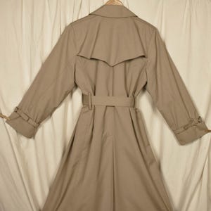 K&ouml;nnte beinhalten: Ein beiger Trenchcoat mit G&uuml;rtel und doppelreihigem Verschluss. Der Mantel hat ein klassisches Design und ist aus leichtem Stoff gefertigt.