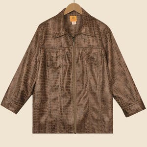 May include: Brown faux-leather jacket with a crocodile skin texture. The jacket features a collared neckline, a zip closure, and two chest pockets. The jacket is hanging on a wooden hanger.