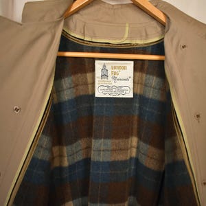 K&ouml;nnte beinhalten: Ein beiger Trenchcoat mit einem karierten Futter. Der Mantel hat einen Rei&szlig;verschluss und Kn&ouml;pfe. Das Futter ist ein braunes, blaues und wei&szlig;es Karomuster. Der Mantel hat ein Etikett mit der Aufschrift "London Fog Maincoats".