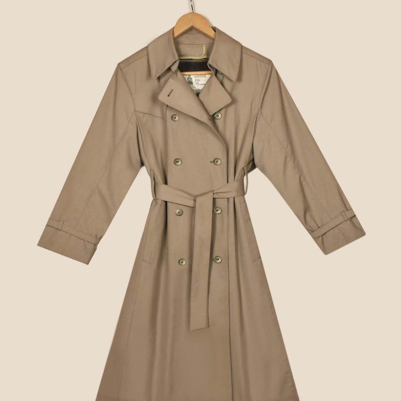 K&ouml;nnte beinhalten: Ein beiger Trenchcoat mit doppelreihigem Verschluss, einem G&uuml;rtel und langen &Auml;rmeln. Der Mantel hat ein klassisches Design und ist aus einem leichten Stoff gefertigt.