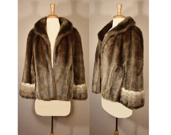 May include: A brown faux fur jacket with a shawl collar and long sleeves. The cuffs are trimmed with a lighter brown fur.