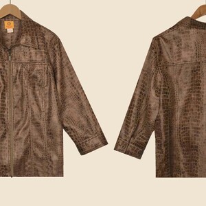 May include: Brown faux leather jacket with a crocodile skin texture, displayed on a wooden hanger. The jacket has a collared neckline, a zip-up front, and two chest pockets. The back of the jacket is also visible.