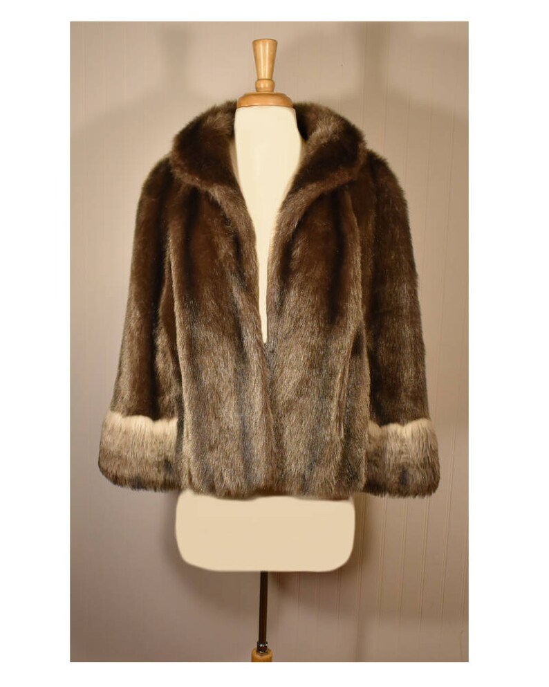 Peut inclure: Une veste en fausse fourrure marron avec une bordure blanche autour du col et des poignets. La veste est ouverte &agrave; l'avant et est expos&eacute;e sur un mannequin.