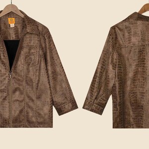 May include: Brown faux-leather jacket with a crocodile skin texture, front and back views. The jacket has a collared neckline, two chest pockets, and a zip-up closure. The sleeves are long, and the jacket hangs on a wooden hanger.